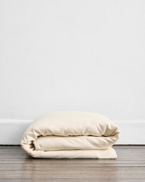The Crème 100% French Flax Linen Duvet Cover by Bed Threads, in a soft cream shade, is neatly folded on a wooden floor against a white wall—ideal for temperature-regulating comfort.