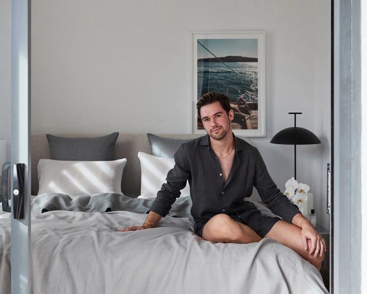 A man in a dark shirt and shorts sits on a neatly made bed with grey and white bedding, in a modern bedroom with neutral decor, pillows, a lamp, and a framed nautical photo on the wall.