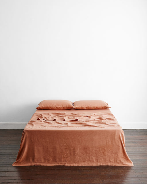 A neatly made bed with two pillows in Bed Threads’ Hazelnut 100% French Flax Linen King Pillowcases is topped with an orange-brown bedspread, centered against a plain white wall on a dark wooden floor.
