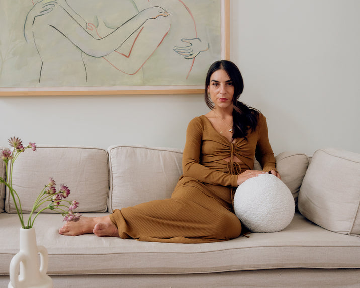 A woman in a brown dress sits barefoot on a beige sofa, leaning against a white textured pillow. A vase with purple flowers is on the table in front of her, and a minimalist line drawing hangs on the wall behind her.