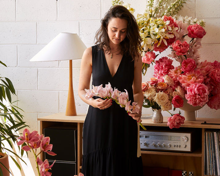 A woman in a black dress stands indoors, holding a stem of pink flowers. She is surrounded by various vibrant flowers and a record player on a wooden shelf, with a white lamp and potted plant nearby.