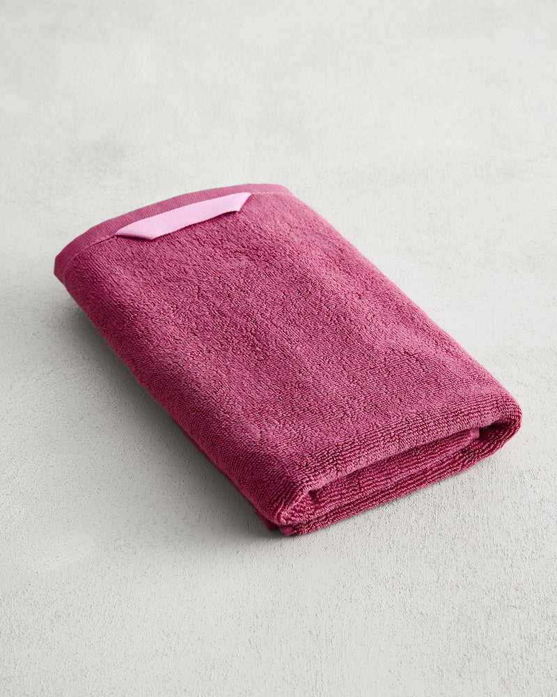 The Berry 100% Cotton Terry Hand Towel by Bed Threads, crafted from plush 650GSM maroon terry and featuring a light pink hanging loop, is displayed on a light gray surface.