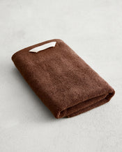 The Bed Threads Espresso 100% Cotton Terry Hand Towel rests on a light gray surface, displaying its plush 650GSM texture and quick-drying features, with a small white tag accenting its rich brown color. - view