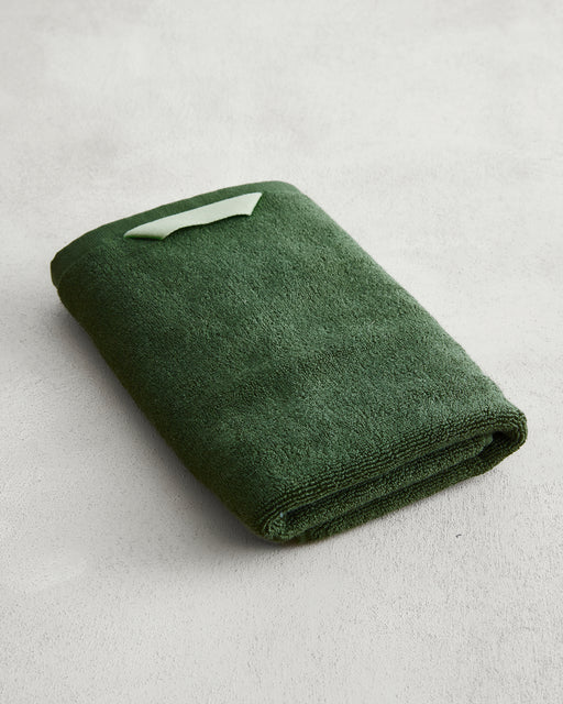The Bed Threads Moss 100% Cotton Terry Hand Towel, in dark green with a light green label, is neatly folded on a light grey textured surface. Crafted from plush 650GSM cotton, it offers softness and exceptional absorbency.
