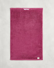 The Bed Threads Berry 100% Cotton Terry Hand Towel is a rectangular, deep pink towel with plush 650GSM texture, a light pink hanging loop at the top, and a small white label in the bottom right corner, shown flat on a textured surface. - view