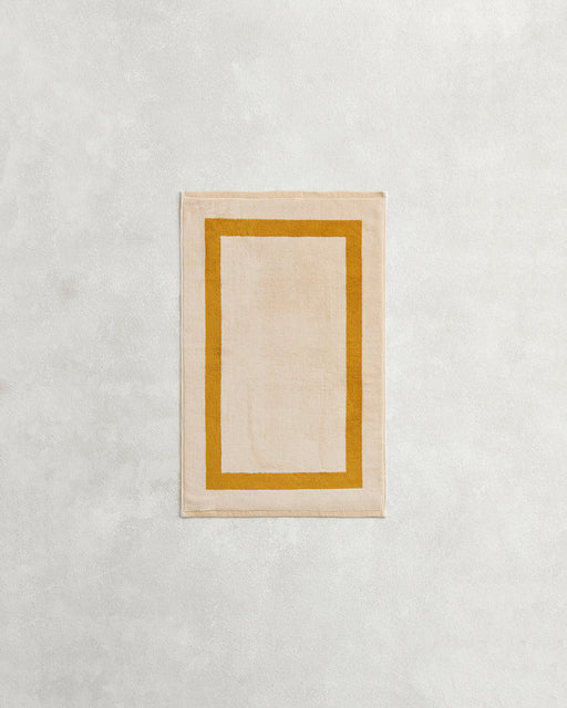 A rectangular beige rug with a mustard border rests on a light grey surface, reflecting the plush 650GSM softness and two-toned style of Bed Threads' Crème & Turmeric 100% Cotton Terry Hand Towel.