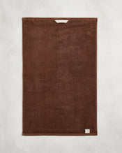 The Bed Threads Espresso 100% Cotton Terry Hand Towel, featuring plush 650GSM brown terry with a small white tag and a white hanging loop, is neatly laid flat on a light gray textured background. - view