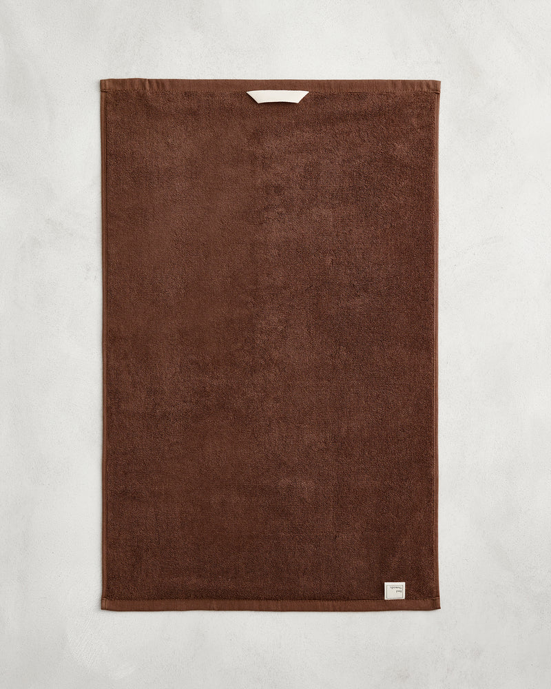 The Bed Threads Espresso 100% Cotton Terry Hand Towel, featuring plush 650GSM brown terry with a small white tag and a white hanging loop, is neatly laid flat on a light gray textured background.