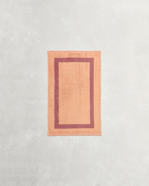 A rectangular peach bath mat with a darker peach border lies on a light gray surface, perfectly complementing Bed Threads’ Terracotta & Pink Clay Two-Toned 100% Cotton Terry Hand Towel.