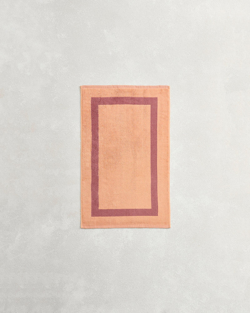 A rectangular peach bath mat with a darker peach border lies on a light gray surface, perfectly complementing Bed Threads’ Terracotta & Pink Clay Two-Toned 100% Cotton Terry Hand Towel.
