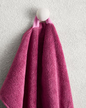The Bed Threads Berry 100% Cotton Terry Hand Towel, in pink, hangs by a loop on a round white hook against a textured light gray wall, offering quick-drying convenience and plush softness. - view
