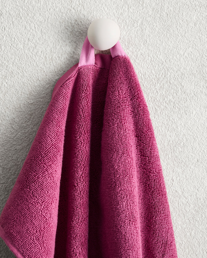 The Bed Threads Berry 100% Cotton Terry Hand Towel, in pink, hangs by a loop on a round white hook against a textured light gray wall, offering quick-drying convenience and plush softness.