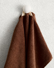 A Bed Threads Espresso 100% Cotton Terry Hand Towel hangs on a round white wall hook against a light, textured wall. - view