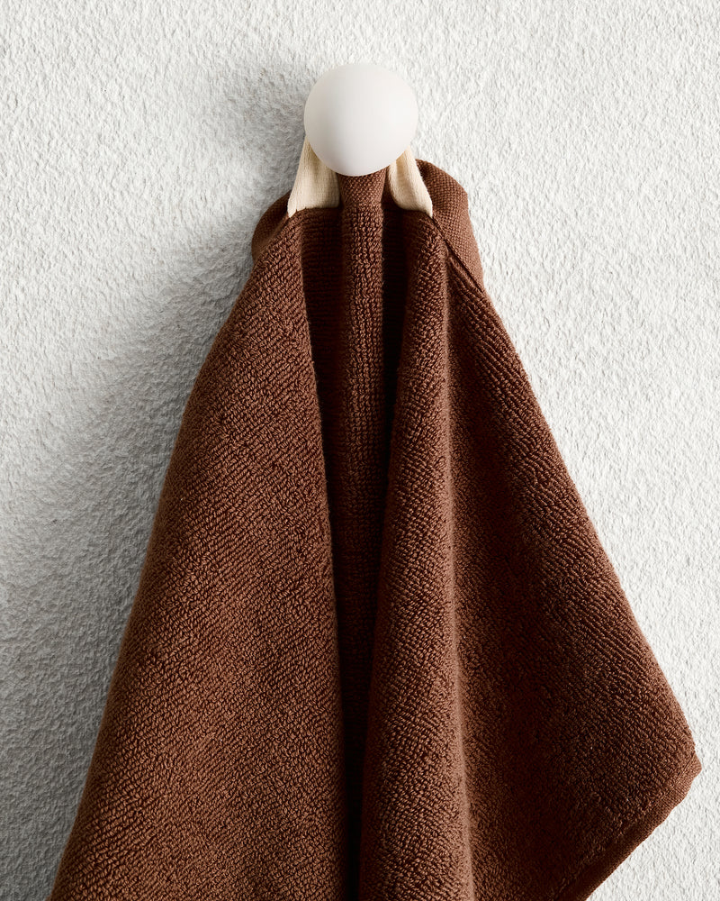 A Bed Threads Espresso 100% Cotton Terry Hand Towel hangs on a round white wall hook against a light, textured wall.