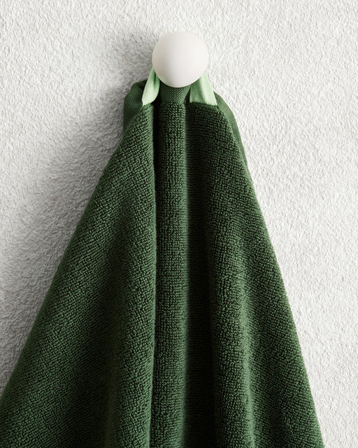 A Moss 100% Cotton Terry Hand Towel from Bed Threads hangs neatly on a round white hook, set against a textured light gray wall.