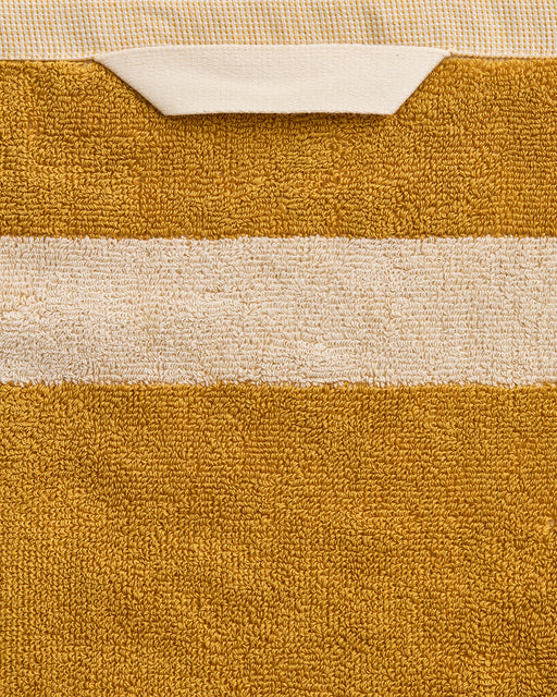 A close-up of the Bed Threads Crème & Turmeric Two-Toned 100% Cotton Terry Hand Towel, made from plush 650GSM terry, featuring a beige stripe and fabric loop, set against a textured background. Comes as a set of two.