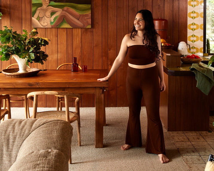 A woman in a brown strapless top and flared pants stands barefoot in a cozy, wood-paneled dining room, smiling and resting one hand on a wooden table. Vintage decor and warm colors fill the room.