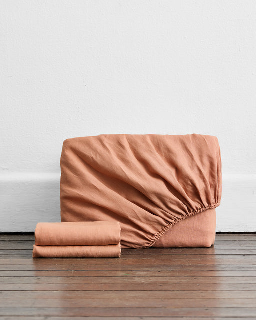 A Hazelnut 100% French Flax Linen Fitted Sheet Set by Bed Threads with two matching pillowcases is folded on a wooden floor, set against a white wall.