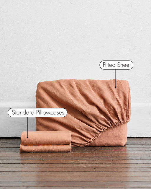 The Bed Threads Hazelnut 100% French Flax Linen Fitted Sheet Set, featuring a rich rust hue, is pictured upright with two matching pillowcases on a wooden floor—each item labeled to highlight this premium linen bedding set.