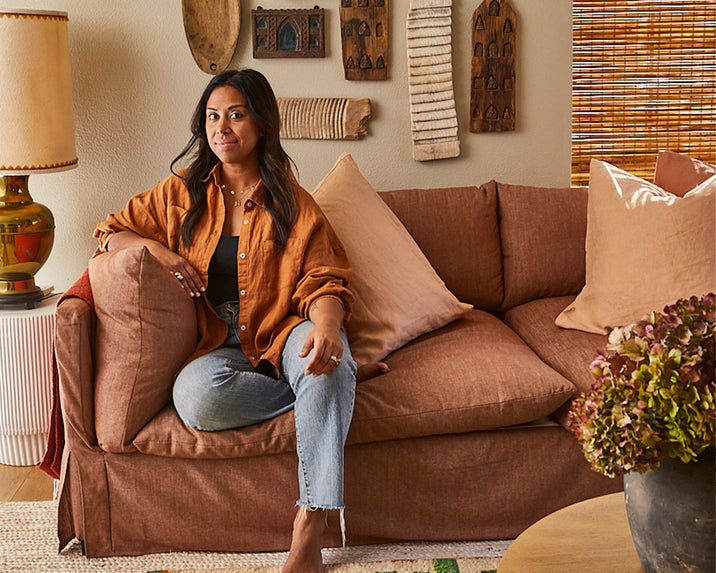 A woman with long dark hair sits barefoot on a brown couch in a cozy, warmly lit living room decorated with textured pillows, rustic wall art, a lamp, and woven blinds.