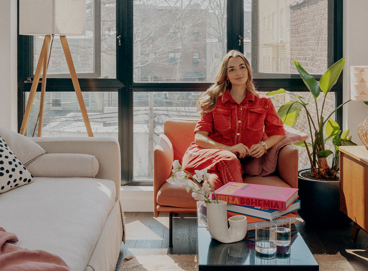 A woman in a red dress sits in an armchair by a large window in a bright, modern living room with plants, books, a lamp, and a coffee table with drinks and flowers.