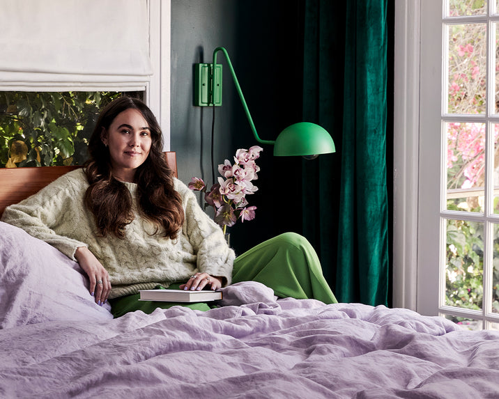 A woman with long brown hair sits on a bed covered in light purple bedding, holding a tablet. She wears a cream sweater and green pants. Behind her is a window with green curtains and a green wall lamp, with flowers nearby.