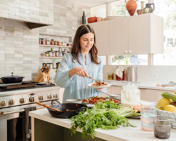 A woman in a light blue shirt stands in a modern kitchen, serving food onto a plate. Fresh vegetables, bowls, and utensils are spread across the counter, and shelves of spices line the wall behind her.