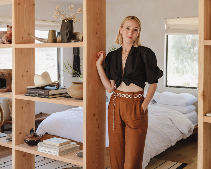 A woman stands by a wooden bookshelf in a cozy bedroom with white bedding. She wears a black top and brown pants, and the room has natural light, plants, and books on shelves.