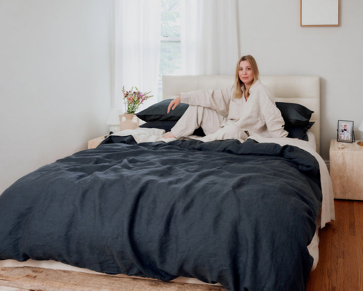 A woman in light pajamas sits on a neatly made bed with dark bedding in a bright, minimal bedroom. There are flowers and a framed photo on a nightstand, with large windows letting in natural light.