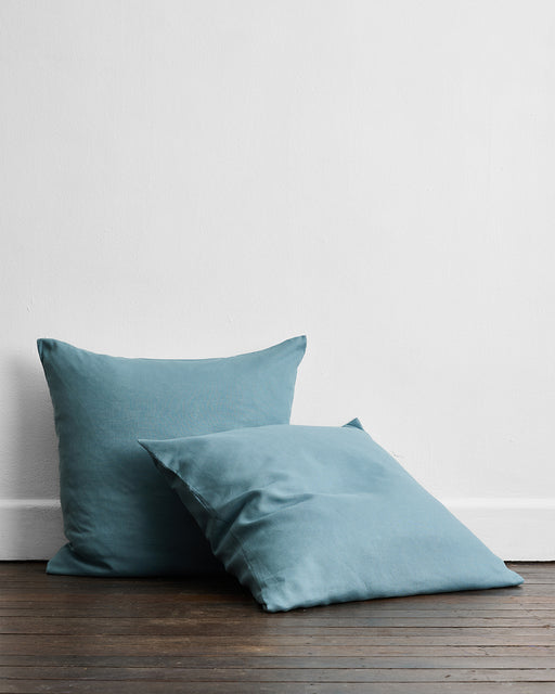 Two Lagoon 100% French Flax Linen European Pillowcases by Bed Threads are placed against a white wall on a wooden floor, with one upright and the other lying in front and slightly overlapping it.