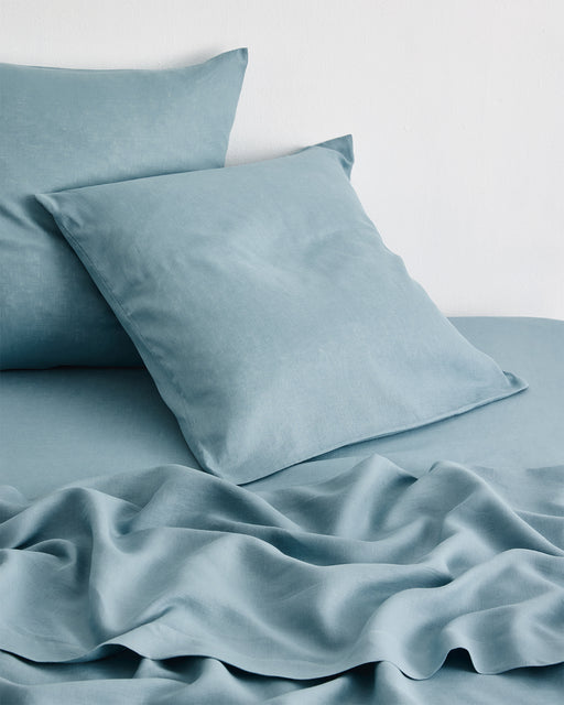 Two Lagoon 100% French Flax Linen European Pillowcases by Bed Threads rest against a matching sheet and blanket on a bed. The lightly rumpled bedding and plain white wall create a soft, inviting scene.