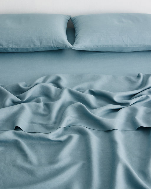 A neatly made bed with two pillows features the Lagoon 100% French Flax Linen Fitted Sheet by Bed Threads in light blue, loosely draped and slightly wrinkled, set against a white background.