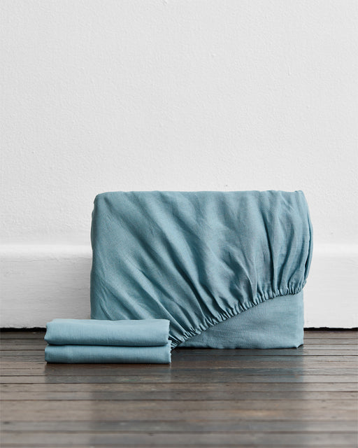 The Lagoon 100% French Flax Linen Fitted Sheet Set by Bed Threads stands upright with two matching pillowcases beside it, adding an elegant touch of blue to your bedding essentials.