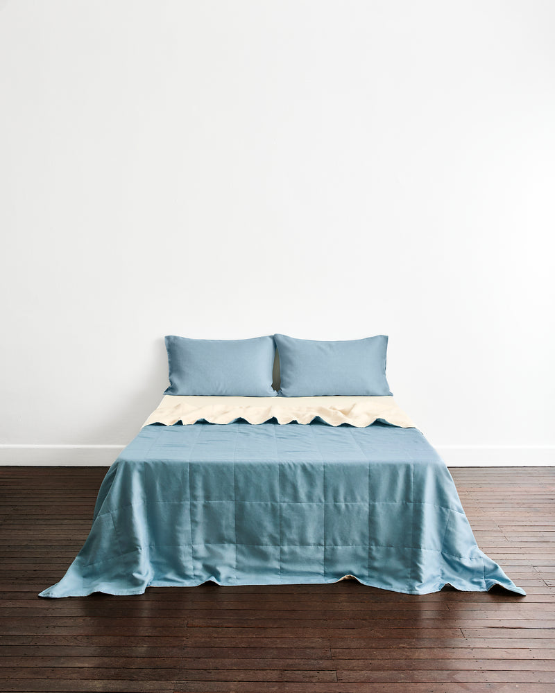 A neatly made bed with a dusty blue quilt cover in Lagoon & Creme and matching pillowcases in Lagoon, set against a plain white wall and dark wood flooring.
