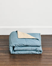 A folded dusty blue quilt cover in Lagoon & Creme with a cream underside, displayed on a dark wooden floor against a white wall. - view