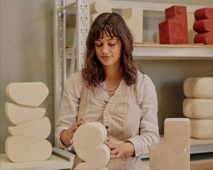 A woman with wavy brown hair works on a cream-colored abstract sculpture in a studio, surrounded by other geometric sculptures and shelves in the background.