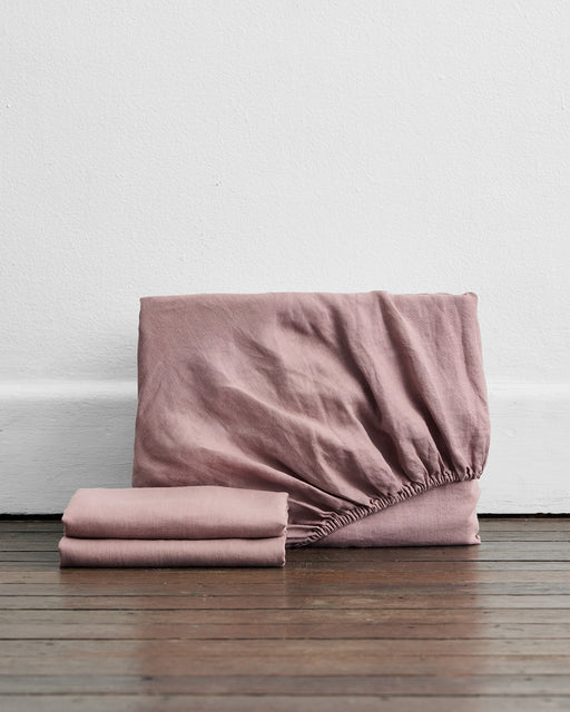 The Bed Threads Lavender 100% French Flax Linen Fitted Sheet Set, featuring a fitted sheet and two flat sheets, is displayed folded on a wooden floor against a white wall.