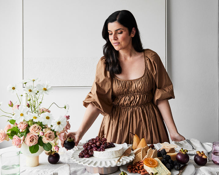 A woman in a brown dress stands at a table with a cheese and fruit platter, arranging food. A vase of pink roses and white daisies sits nearby in a bright, minimalist room.