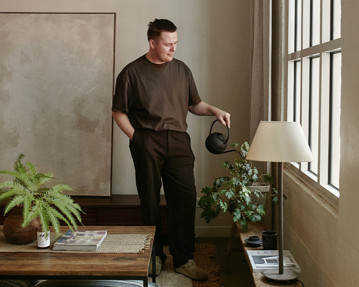 A person stands by a window in a cozy, sunlit room, holding a black watering can and tending to a potted plant beside a table with magazines, a lamp, and another plant. The room has neutral tones and minimalist decor.