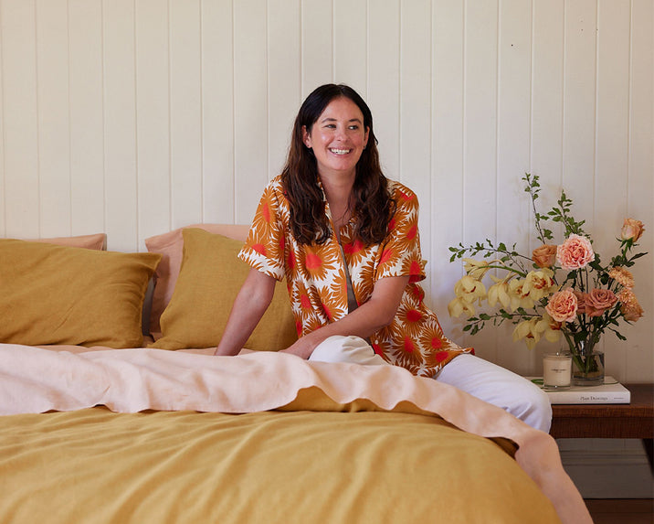A woman with long brown hair, wearing a bright floral shirt and white pants, sits smiling on a bed with mustard yellow linens. A wooden bedside table holds a vase of flowers, a book, and a candle.