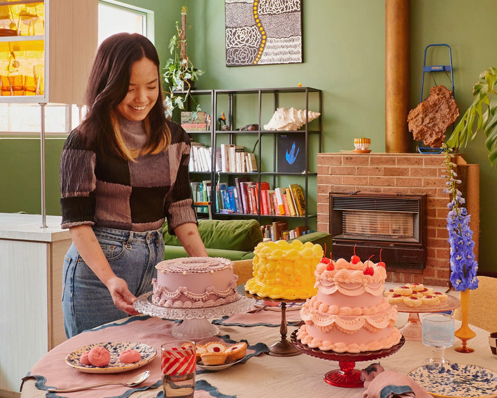 A woman smiles as she sets a lavender cake on a table with more decorated cakes, pastries, and drinks. The cozy room features green walls, bookshelves, art, and a brick fireplace in the background.