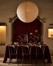 A warmly lit dining table set for four with dark wooden chairs, a burgundy tablecloth, candles, and flower arrangements. A large round paper lantern hangs above, and the background features rustic stone walls and a red arched door. - view