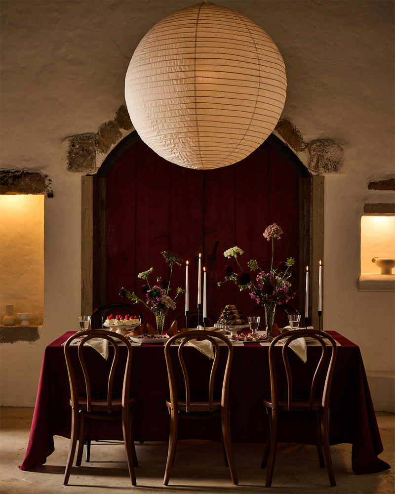A warmly lit dining table set for four with dark wooden chairs, a burgundy tablecloth, candles, and flower arrangements. A large round paper lantern hangs above, and the background features rustic stone walls and a red arched door.