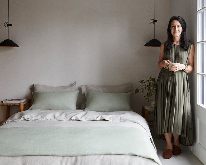 A woman in a long, pleated dress stands by a neatly made bed with light green and beige bedding in a minimal, softly lit bedroom, holding a cup and smiling gently at the camera.