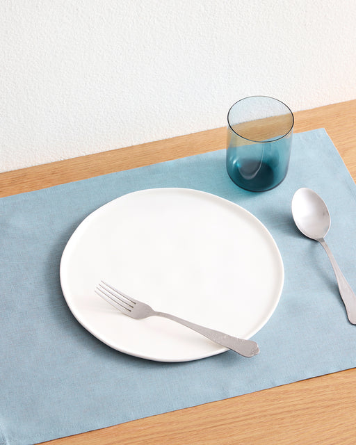 A white plate with a fork sits on a Lagoon 100% French Flax Linen Placemat by Bed Threads atop a wooden table, next to a spoon and blue-tinted glass tumbler.