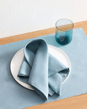 A Lagoon 100% French Flax Linen Napkin by Bed Threads is folded on a white plate atop a matching placemat on a wooden table, with a translucent blue glass nearby. - view
