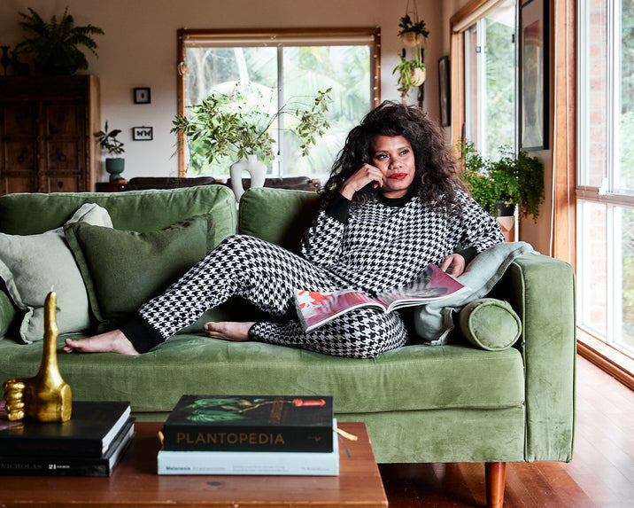 A woman in a black and white houndstooth outfit lounges on a green velvet couch, reading a magazine. The room is filled with houseplants and natural light from large windows. A coffee table with books is in the foreground.