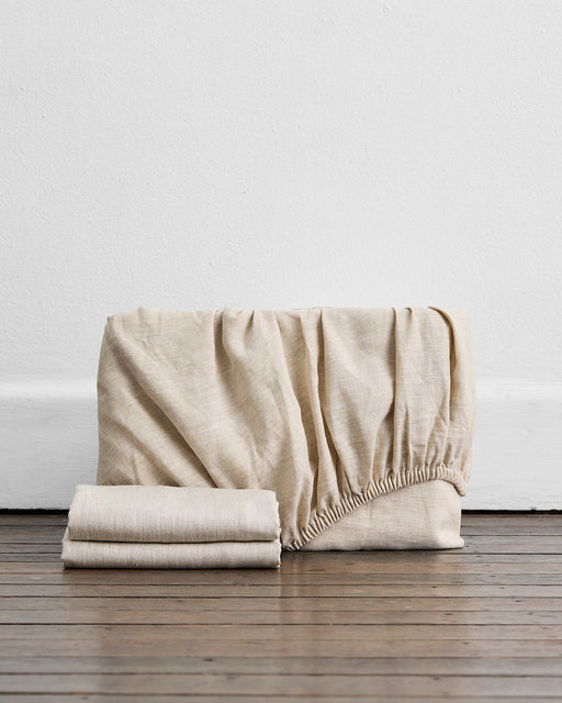The Bed Threads Oatmeal 100% French Flax Linen Fitted Sheet Set, featuring elastic edges and a folded sheet, is shown on a wooden floor against a white wall.