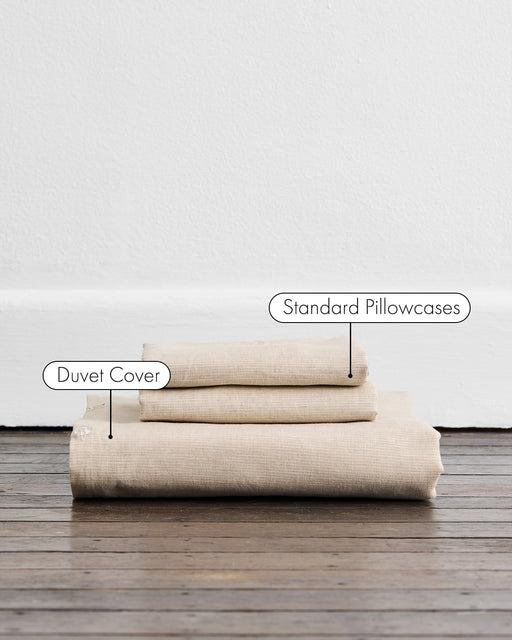Three neatly folded Oatmeal & White Stripe 100% French Flax Linen pieces by Bed Threads—a Duvet Cover (bottom) and two Standard Pillowcases—are displayed on a wooden floor against a white wall.