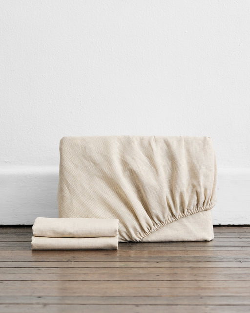 The Bed Threads Oatmeal & White Stripe 100% French Flax Linen Fitted Sheet Set rests on a wooden floor by a white wall, with one folded fitted sheet and two flat sheets—ideal for elevating your linen bedding collection.